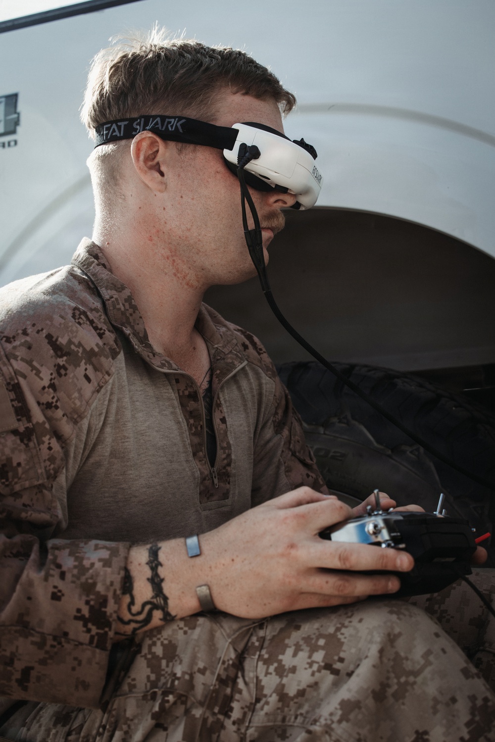 A military service member wearing USMC MARPAT desert camouflage uniform is operating an FPV (First Person View) drone using a handheld radio controller. He is wearing Fat Shark FPV goggles, which provide a live video feed from the drone's camera. The setting appears to be near a military vehicle or shelter structure.