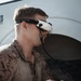 A military service member wearing USMC MARPAT desert camouflage uniform is operating an FPV (First Person View) drone using a handheld radio controller. He is wearing Fat Shark FPV goggles, which provide a live video feed from the drone's camera. The setting appears to be near a military vehicle or shelter structure.