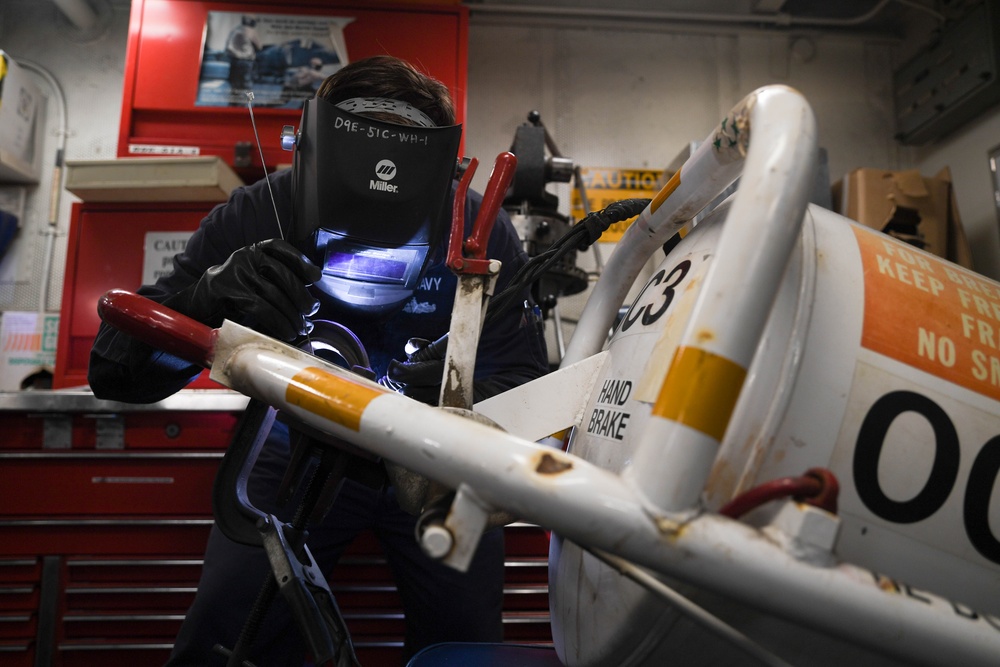 A welder wearing a Miller brand auto-darkening welding helmet is actively welding a metal component, likely part of a piece of military or industrial equipment. The work is being performed in what appears to be a workshop or maintenance bay, with red tool cabinets visible in the background. The welder's helmet is marked 'D9E-51C-WH-1' and the uniform appears to bear the word 'NAVY', suggesting a U.S. Navy maintenance environment.