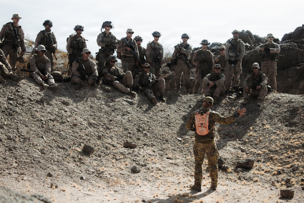 A military instructor or observer wearing an orange safety vest marked '3D' addresses a group of approximately 15-20 soldiers positioned on a rocky desert hillside. The soldiers are equipped with full combat gear, weapons, and helmets, suggesting this is a training exercise or after-action review. The arid, rocky terrain is consistent with a desert training environment such as those found in the American Southwest.
