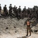 A military instructor or observer wearing an orange safety vest marked '3D' addresses a group of approximately 15-20 soldiers positioned on a rocky desert hillside. The soldiers are equipped with full combat gear, weapons, and helmets, suggesting this is a training exercise or after-action review. The arid, rocky terrain is consistent with a desert training environment such as those found in the American Southwest.