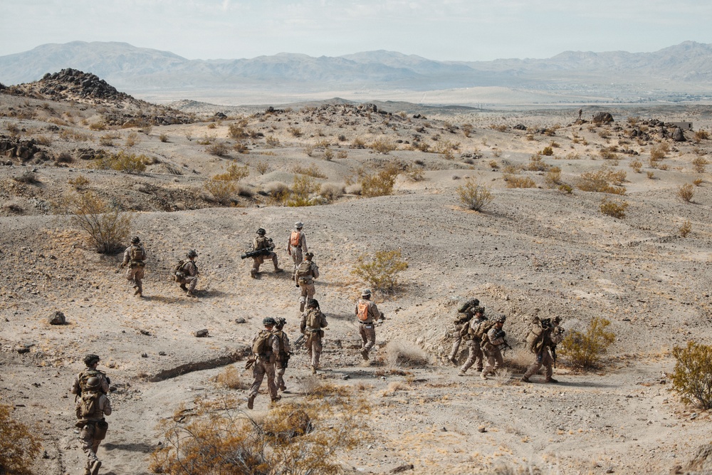 A group of military personnel in desert camouflage uniforms are conducting a patrol or training exercise across an arid, rocky desert landscape. Several individuals wearing orange safety vests are visible among the group, suggesting this is a supervised training exercise rather than a combat operation. The terrain features sparse desert vegetation, rocky outcroppings, and distant mountain ranges consistent with a Southwestern U.S. training environment.