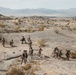 A group of military personnel in desert camouflage uniforms are conducting a patrol or training exercise across an arid, rocky desert landscape. Several individuals wearing orange safety vests are visible among the group, suggesting this is a supervised training exercise rather than a combat operation. The terrain features sparse desert vegetation, rocky outcroppings, and distant mountain ranges consistent with a Southwestern U.S. training environment.