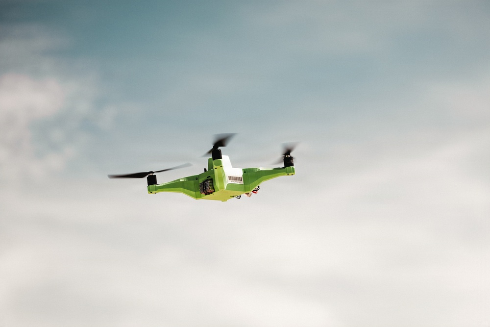 A bright green quadcopter drone is captured mid-flight against a partly cloudy blue-grey sky. The drone appears to be carrying a small white package or payload on top of its body, suggesting a delivery or transport function. The rotors are visibly blurred due to their spinning motion, indicating active flight.