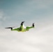A bright green quadcopter drone is captured mid-flight against a partly cloudy blue-grey sky. The drone appears to be carrying a small white package or payload on top of its body, suggesting a delivery or transport function. The rotors are visibly blurred due to their spinning motion, indicating active flight.
