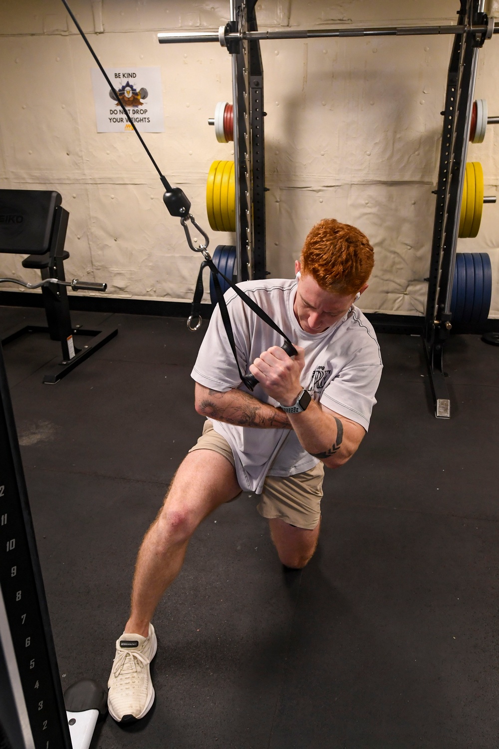A red-haired man is performing a cable machine exercise in a gym, appearing to do a kneeling cable row or pull exercise while in a lunge position. He is wearing a white t-shirt, khaki shorts, and white sneakers, with visible tattoos on his forearms and a smartwatch on his wrist. The gym setting includes a power rack with loaded barbells, a weight bench, and a motivational sign on the wall.