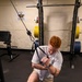 A red-haired man is performing a cable machine exercise in a gym, appearing to do a kneeling cable row or pull exercise while in a lunge position. He is wearing a white t-shirt, khaki shorts, and white sneakers, with visible tattoos on his forearms and a smartwatch on his wrist. The gym setting includes a power rack with loaded barbells, a weight bench, and a motivational sign on the wall.