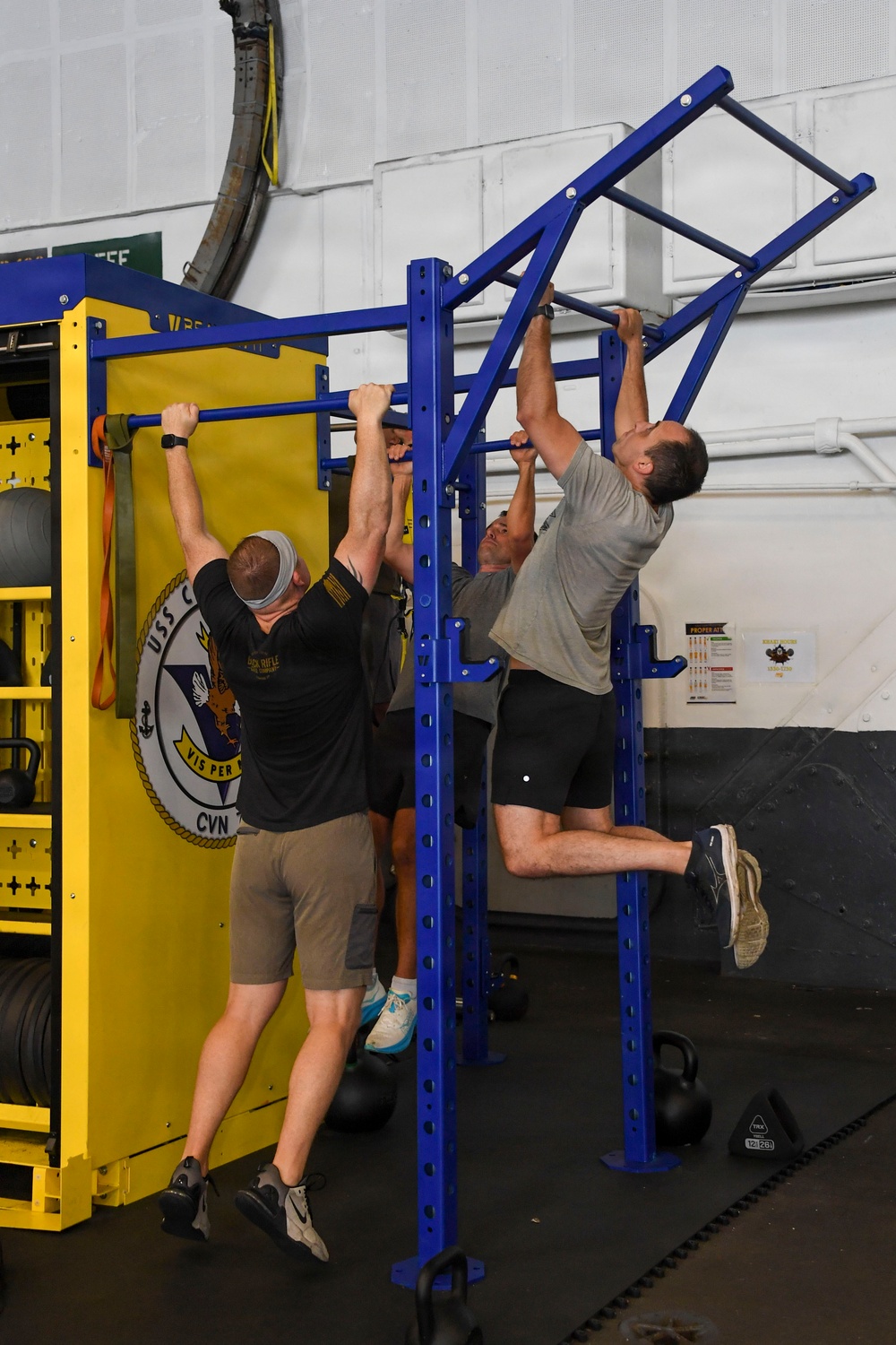 Multiple individuals are performing pull-ups on a blue metal pull-up rig in what appears to be a military fitness facility. A yellow equipment rack bearing a USS CVN ship insignia with the motto 'VIS PER' is visible in the background. The setting is an indoor gym with kettlebells, resistance bands, and fitness posters on the walls.