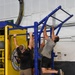 Multiple individuals are performing pull-ups on a blue metal pull-up rig in what appears to be a military fitness facility. A yellow equipment rack bearing a USS CVN ship insignia with the motto 'VIS PER' is visible in the background. The setting is an indoor gym with kettlebells, resistance bands, and fitness posters on the walls.