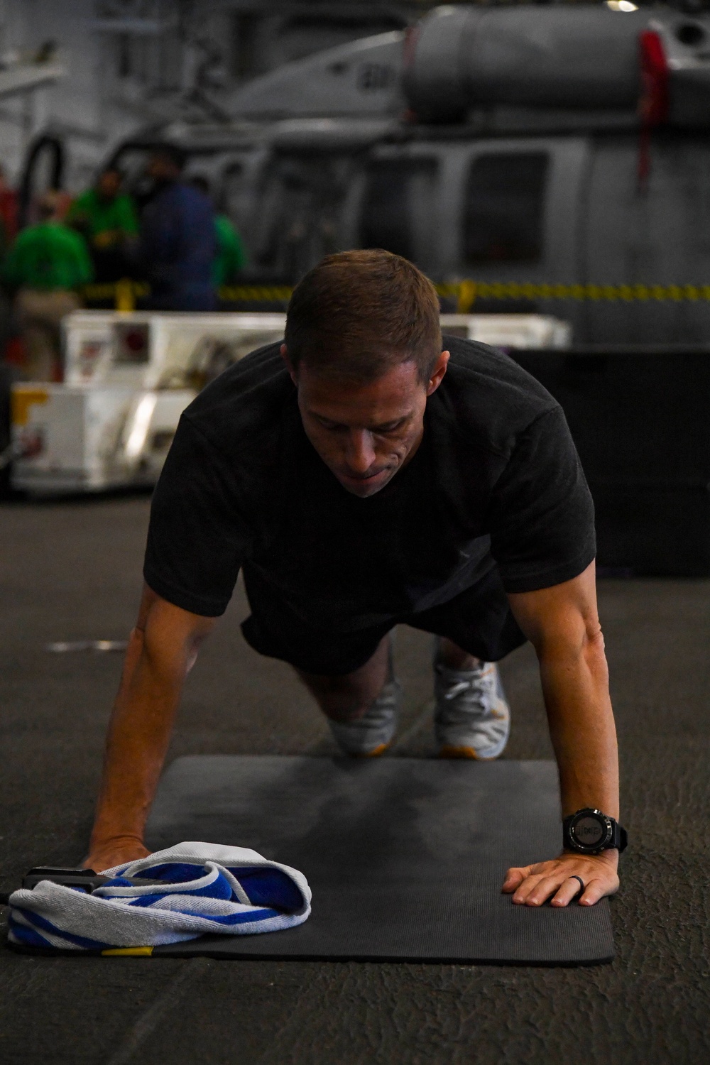 A man in a black t-shirt is performing push-ups on a dark exercise mat in what appears to be the hangar bay of a naval vessel. He is wearing athletic shoes and a black sports watch, with a folded towel and what appears to be a phone placed on the mat beside him. In the background, military equipment, vehicles, and personnel in colored jerseys are visible.