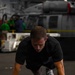 A man in a black t-shirt is performing push-ups on a dark exercise mat in what appears to be the hangar bay of a naval vessel. He is wearing athletic shoes and a black sports watch, with a folded towel and what appears to be a phone placed on the mat beside him. In the background, military equipment, vehicles, and personnel in colored jerseys are visible.