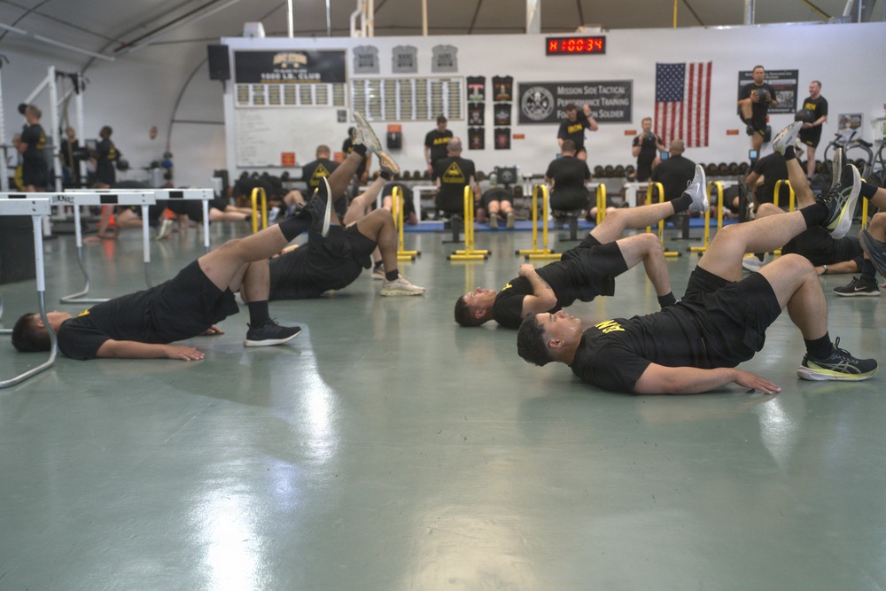 A large group of military personnel in matching black Army PT (Physical Training) uniforms are performing synchronized floor exercises in a well-equipped gymnasium. The foreground shows soldiers executing single-leg hip bridge exercises on a green-painted floor, while additional soldiers and equipment are visible in the background. A digital timer displaying '8:00:34' is visible on the wall, indicating a timed training session.
