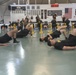 A large group of military personnel in matching black Army PT (Physical Training) uniforms are performing synchronized floor exercises in a well-equipped gymnasium. The foreground shows soldiers executing single-leg hip bridge exercises on a green-painted floor, while additional soldiers and equipment are visible in the background. A digital timer displaying '8:00:34' is visible on the wall, indicating a timed training session.
