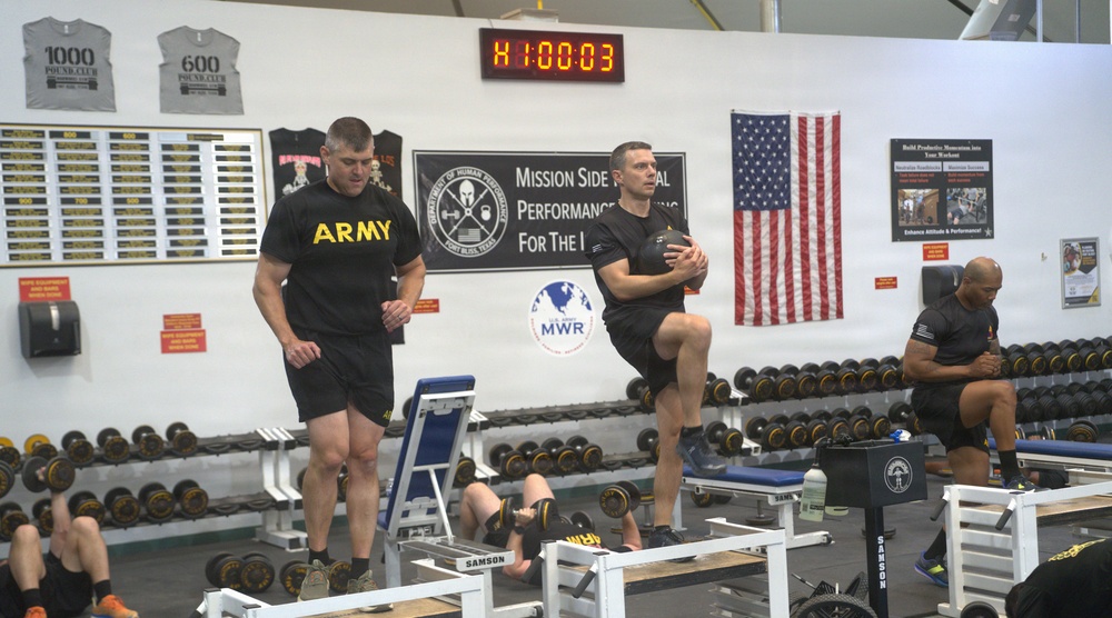 U.S. Army soldiers are exercising in a military fitness facility, likely at Fort Bliss, Texas, based on visible signage. The gym features a 'Department of Human Performance' banner, U.S. Army MWR (Morale, Welfare, and Recreation) branding, and an American flag on the wall. A digital timer reads 1:00:03, suggesting a timed workout session is in progress.