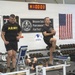 U.S. Army soldiers are exercising in a military fitness facility, likely at Fort Bliss, Texas, based on visible signage. The gym features a 'Department of Human Performance' banner, U.S. Army MWR (Morale, Welfare, and Recreation) branding, and an American flag on the wall. A digital timer reads 1:00:03, suggesting a timed workout session is in progress.