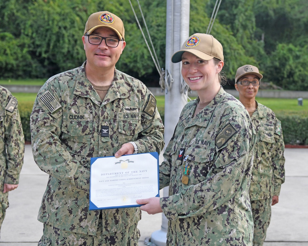 A U.S. Navy officer named Cudnik is presenting a Department of the Navy certificate, specifically a Navy and Marine Corps Achievement Medal, to a female sailor during a formal award ceremony. Both individuals are wearing Navy Working Uniform (NWU Type III) camouflage and tan unit caps. A third service member is visible in the background observing the ceremony.