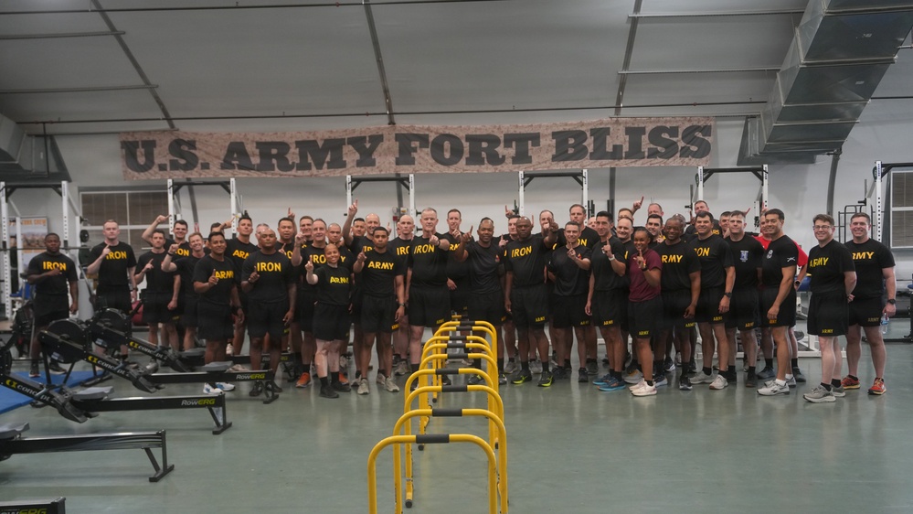 A large group of U.S. Army personnel pose for a group photo inside a fitness facility at Fort Bliss. The individuals are wearing black Army physical training uniforms with 'ARMY' and 'IRON' text, and many are giving thumbs-up gestures. The setting is a well-equipped gymnasium with a prominent 'U.S. ARMY FORT BLISS' banner on the wall behind them.