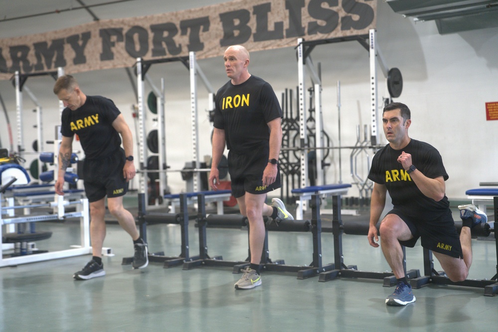 Three individuals are exercising in a military gym at Fort Bliss, as indicated by the large sign on the wall. Two are wearing black 'ARMY' branded shirts and shorts, while the center individual wears a black 'IRON' shirt. They appear to be performing lower-body exercises, including lunges and stepping movements.
