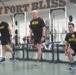 Three individuals are exercising in a military gym at Fort Bliss, as indicated by the large sign on the wall. Two are wearing black 'ARMY' branded shirts and shorts, while the center individual wears a black 'IRON' shirt. They appear to be performing lower-body exercises, including lunges and stepping movements.