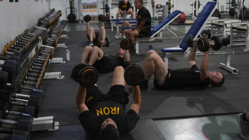 A group of individuals wearing black Army-branded athletic uniforms are performing dumbbell floor press exercises in a military gym facility. Multiple personnel are lying on the floor pressing dumbbells simultaneously, while others are seated or resting in the background. The gym is well-equipped with a full rack of dumbbells along the left wall and various weight training equipment.