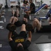 A group of individuals wearing black Army-branded athletic uniforms are performing dumbbell floor press exercises in a military gym facility. Multiple personnel are lying on the floor pressing dumbbells simultaneously, while others are seated or resting in the background. The gym is well-equipped with a full rack of dumbbells along the left wall and various weight training equipment.