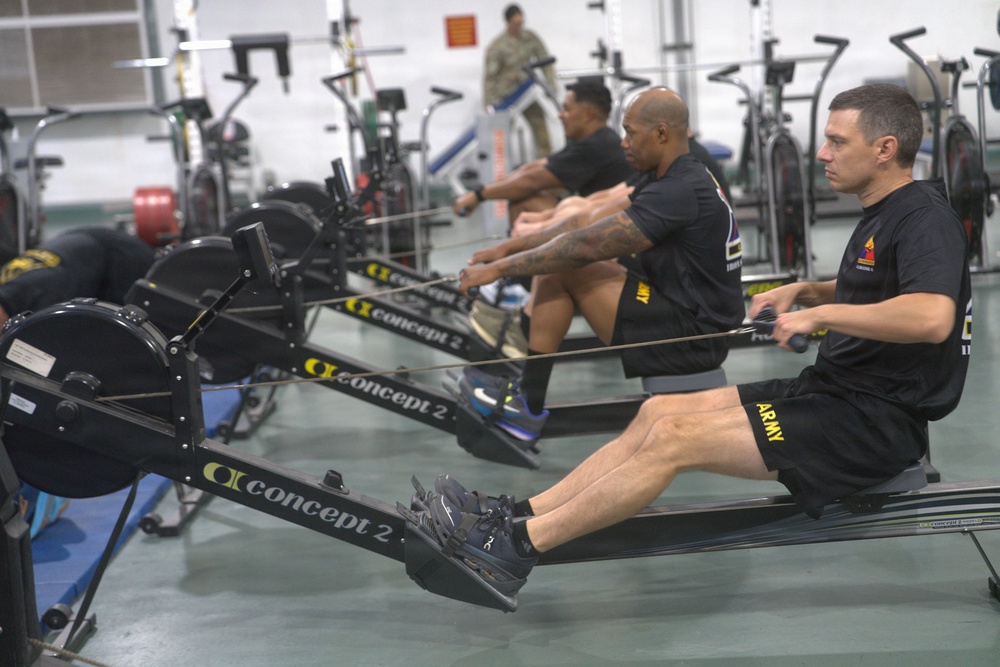 A group of U.S. Army soldiers are using Concept 2 rowing machines in a military fitness facility. Multiple individuals wearing black Army-branded athletic gear are performing synchronized rowing exercises. The gym setting includes additional cardio equipment visible in the background.