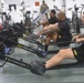 A group of U.S. Army soldiers are using Concept 2 rowing machines in a military fitness facility. Multiple individuals wearing black Army-branded athletic gear are performing synchronized rowing exercises. The gym setting includes additional cardio equipment visible in the background.