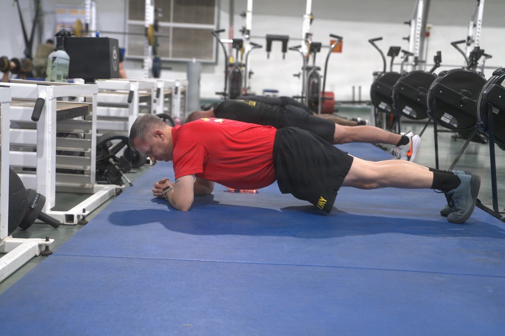 Multiple individuals are performing plank exercises on blue gym mats in a well-equipped fitness facility. The foreground subject is wearing a red shirt and black shorts with 'ARMY' visible in yellow lettering, suggesting a military fitness context. Several rowing machines and other gym equipment are visible in the background.