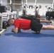 Multiple individuals are performing plank exercises on blue gym mats in a well-equipped fitness facility. The foreground subject is wearing a red shirt and black shorts with 'ARMY' visible in yellow lettering, suggesting a military fitness context. Several rowing machines and other gym equipment are visible in the background.