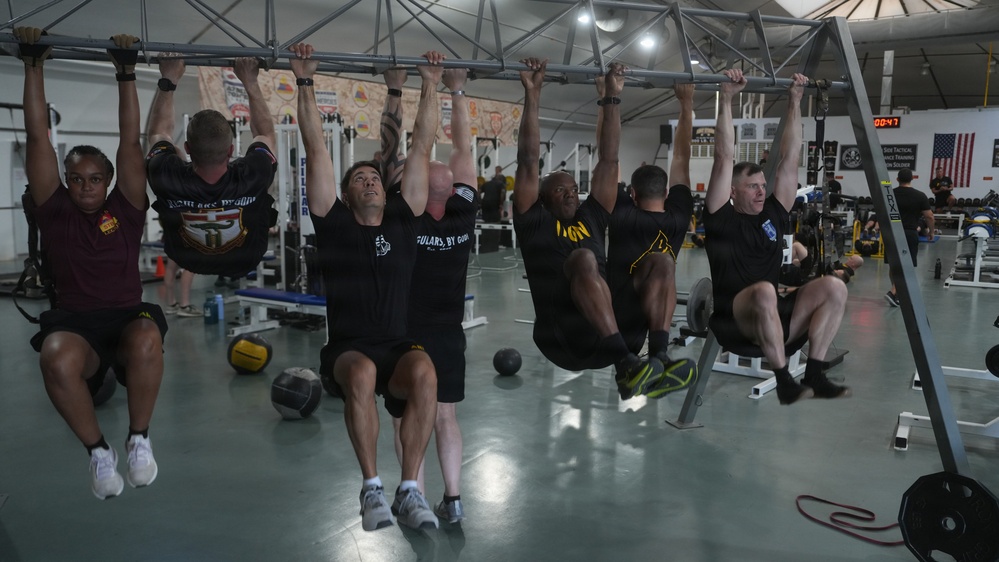 A group of approximately six individuals are performing hanging leg raises or knee tucks simultaneously on a shared overhead pull-up bar structure in a military-style gymnasium. The participants, wearing military physical training attire including black and maroon shirts, are suspended from the bar with their legs raised. The gym is well-equipped with various exercise machines, medicine balls, and motivational signage visible in the background.