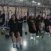 A group of approximately six individuals are performing hanging leg raises or knee tucks simultaneously on a shared overhead pull-up bar structure in a military-style gymnasium. The participants, wearing military physical training attire including black and maroon shirts, are suspended from the bar with their legs raised. The gym is well-equipped with various exercise machines, medicine balls, and motivational signage visible in the background.