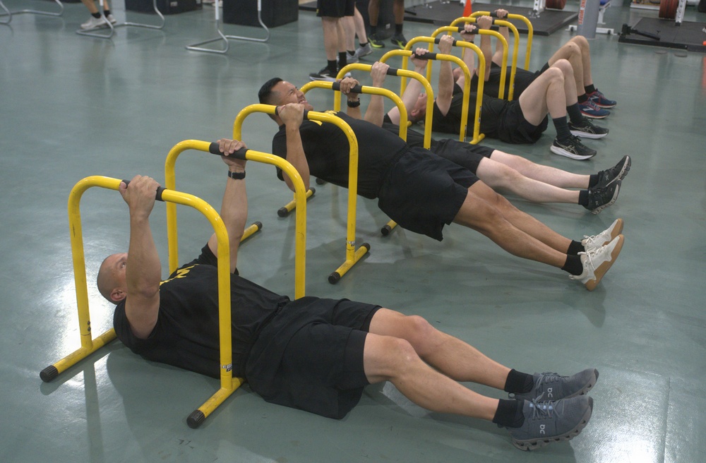A group of individuals dressed in matching black t-shirts and black shorts are performing row exercises using yellow portable parallette bars on a gymnasium floor. They are lying on their backs and pulling their chests up to the bars in a synchronized manner, suggesting a structured physical training session. The setting appears to be a military or institutional fitness facility.