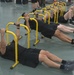A group of individuals dressed in matching black t-shirts and black shorts are performing row exercises using yellow portable parallette bars on a gymnasium floor. They are lying on their backs and pulling their chests up to the bars in a synchronized manner, suggesting a structured physical training session. The setting appears to be a military or institutional fitness facility.