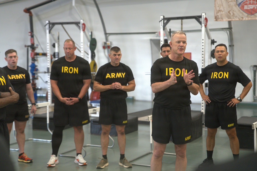 A group of U.S. Army personnel are gathered inside a gym or fitness facility, wearing matching black physical training uniforms with yellow 'ARMY' and 'IRON' text. One individual in the foreground appears to be speaking or briefing the group, gesturing with his hands while others stand attentively. The setting appears to be a military fitness or strength training facility.