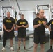 A group of U.S. Army personnel are gathered inside a gym or fitness facility, wearing matching black physical training uniforms with yellow 'ARMY' and 'IRON' text. One individual in the foreground appears to be speaking or briefing the group, gesturing with his hands while others stand attentively. The setting appears to be a military fitness or strength training facility.