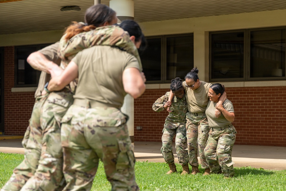 A group of female military personnel in camouflage uniforms are engaged in what appears to be combat training or tactical exercises on a grassy area outside a brick building. Two distinct pairs or groups are visible: one group in the foreground grappling or embracing, and another group in the background where two soldiers appear to be assisting or restraining a third. The setting suggests a military installation or training facility.