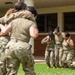 A group of female military personnel in camouflage uniforms are engaged in what appears to be combat training or tactical exercises on a grassy area outside a brick building. Two distinct pairs or groups are visible: one group in the foreground grappling or embracing, and another group in the background where two soldiers appear to be assisting or restraining a third. The setting suggests a military installation or training facility.