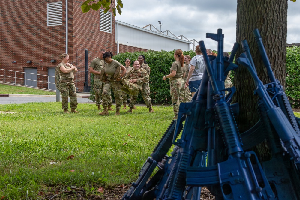A group of military personnel in camouflage uniforms are engaged in what appears to be a combatives or hand-to-hand combat training exercise on a grass area near a brick building. In the foreground, multiple blue training rifles are stacked in a teepee formation against a tree, indicating these are inert training weapons. Several soldiers observe while two or three actively practice a grappling or casualty carry technique.