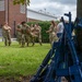 A group of military personnel in camouflage uniforms are engaged in what appears to be a combatives or hand-to-hand combat training exercise on a grass area near a brick building. In the foreground, multiple blue training rifles are stacked in a teepee formation against a tree, indicating these are inert training weapons. Several soldiers observe while two or three actively practice a grappling or casualty carry technique.