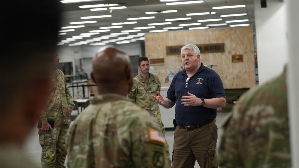 A civilian in a navy blue polo shirt appears to be briefing or addressing a group of military personnel in camouflage uniforms inside what appears to be a military facility or training center. The civilian is gesturing with his hands, suggesting he is actively speaking or presenting information. Several soldiers in OCP (Operational Camouflage Pattern) uniforms are listening, with their backs partially to the camera.