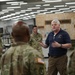 A civilian in a navy blue polo shirt appears to be briefing or addressing a group of military personnel in camouflage uniforms inside what appears to be a military facility or training center. The civilian is gesturing with his hands, suggesting he is actively speaking or presenting information. Several soldiers in OCP (Operational Camouflage Pattern) uniforms are listening, with their backs partially to the camera.