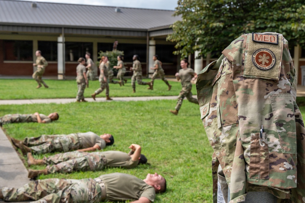 Military personnel in OCP (Operational Camouflage Pattern) uniforms are engaged in what appears to be a medical training exercise outdoors. Some personnel are lying on the grass simulating casualties while others run in the background, likely acting as responders. A prominent uniform in the foreground displays a 'MED' tab and a 'Nationally Registered Emergency Medical Technician' patch.