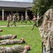 Military personnel in OCP (Operational Camouflage Pattern) uniforms are engaged in what appears to be a medical training exercise outdoors. Some personnel are lying on the grass simulating casualties while others run in the background, likely acting as responders. A prominent uniform in the foreground displays a 'MED' tab and a 'Nationally Registered Emergency Medical Technician' patch.