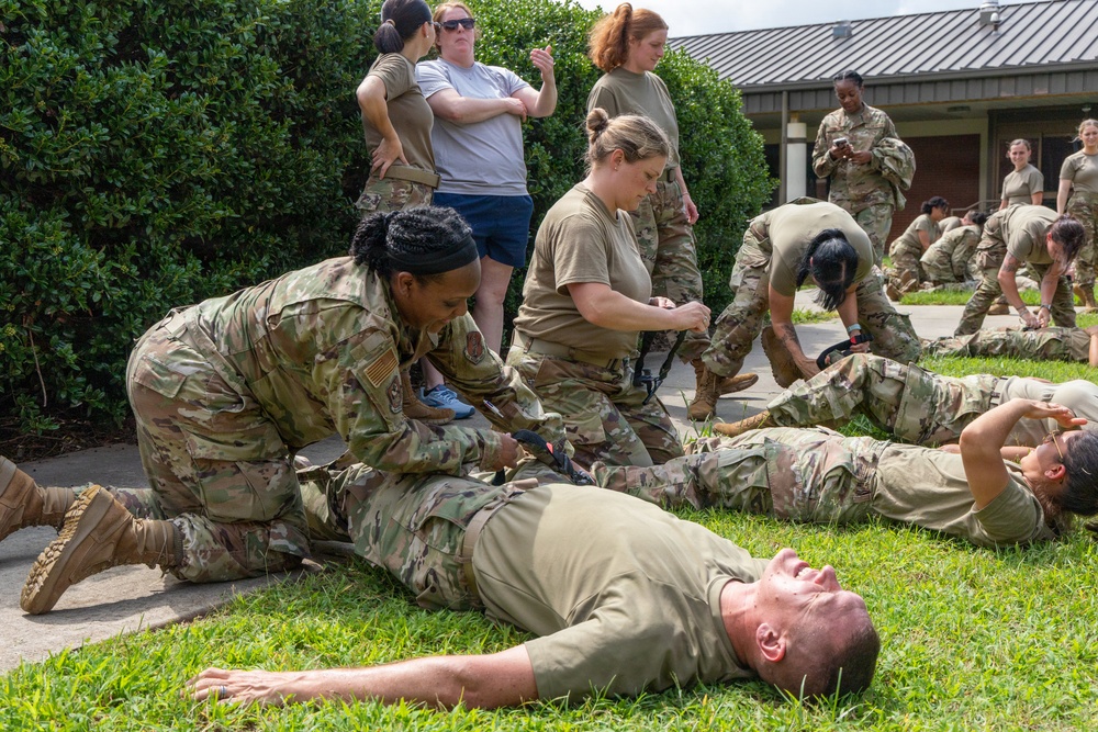 Military personnel in OCP (Operational Camouflage Pattern) uniforms are conducting a tactical combat casualty care (TCCC) or first aid training exercise outdoors. Several soldiers are practicing medical procedures on simulated casualties lying on the ground. The training appears to involve tourniquet application and other emergency medical response techniques.