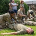 Military personnel in OCP (Operational Camouflage Pattern) uniforms are conducting a tactical combat casualty care (TCCC) or first aid training exercise outdoors. Several soldiers are practicing medical procedures on simulated casualties lying on the ground. The training appears to involve tourniquet application and other emergency medical response techniques.