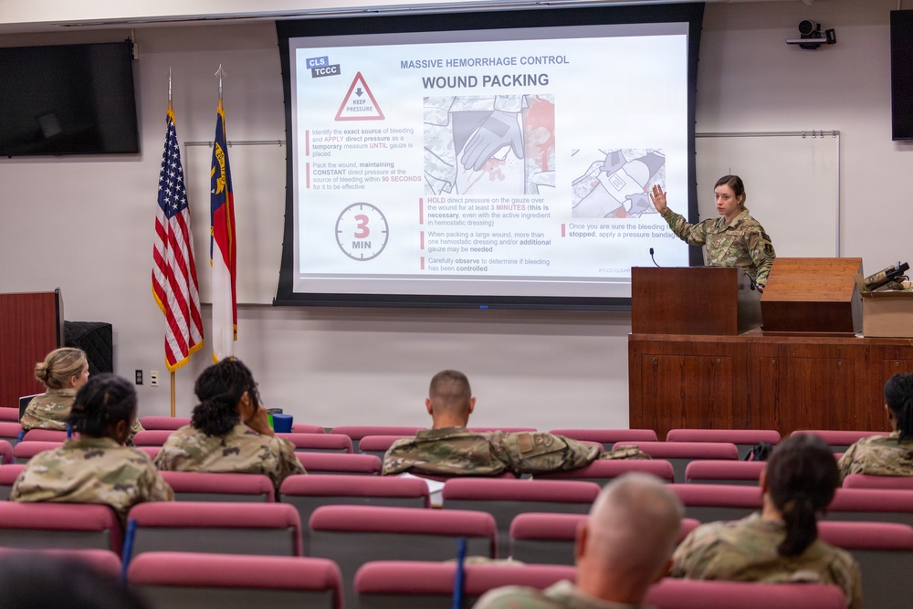 A military service member in camouflage uniform is presenting a medical training briefing on 'Massive Hemorrhage Control - Wound Packing' to an audience of fellow soldiers in a classroom/auditorium setting. The presentation covers Tactical Combat Casualty Care (TCCC) and Combat Lifesaver (CLS) protocols for wound packing techniques. The audience is seated in tiered red theater-style seating facing the instructor and projection screen.