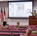 A military service member in camouflage uniform is presenting a medical training briefing on 'Massive Hemorrhage Control - Wound Packing' to an audience of fellow soldiers in a classroom/auditorium setting. The presentation covers Tactical Combat Casualty Care (TCCC) and Combat Lifesaver (CLS) protocols for wound packing techniques. The audience is seated in tiered red theater-style seating facing the instructor and projection screen.