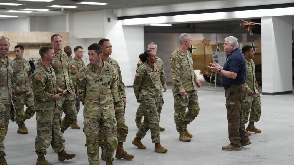 A group of U.S. Army soldiers in OCP (Operational Camouflage Pattern) uniforms are being led on what appears to be a guided tour of a military museum or exhibit facility. A civilian guide in a blue polo shirt is speaking and gesturing to the group as they walk through a large indoor space. Military vehicles and display boards are visible in the background.