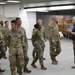 A group of U.S. Army soldiers in OCP (Operational Camouflage Pattern) uniforms are being led on what appears to be a guided tour of a military museum or exhibit facility. A civilian guide in a blue polo shirt is speaking and gesturing to the group as they walk through a large indoor space. Military vehicles and display boards are visible in the background.