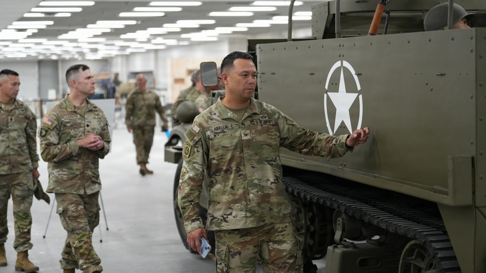 A group of U.S. Army soldiers in MultiCam (OCP) uniforms are touring what appears to be a military museum or exhibit hall. The central figure, whose name tape reads 'ABELLA,' is standing next to a vintage WWII-era half-track vehicle bearing the white Allied star insignia. The setting is an indoor facility with modern lighting, suggesting an organized tour or official visit.